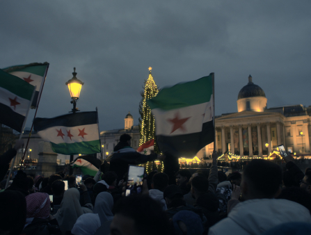 A nighttime protest shot with people holding Syrian flags in front of a government buildling