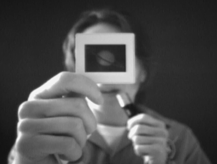 Black and white image of a white man in a shirt holding a microphone in one hand and a slide of Saturn over his face with the other