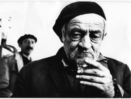 Black and white close-up shot of an aging white man with a beret holding hand up with a cigarette, a man in the background looking on