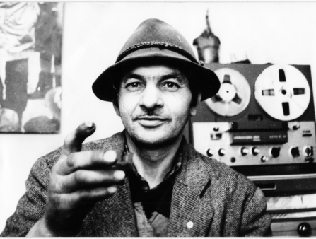 Black and white shot of a white man in a blazer and a fedora holding up his hand up to the camera, with a recording device behind him