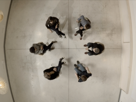 Shot from above of six people sitting in a circle facing one another against a white-tiled floor