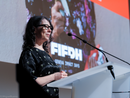 A middle-aged white woman with shoulder-length black wavy hair dons glasses and a black dress with white flowers at a podium with a mic
