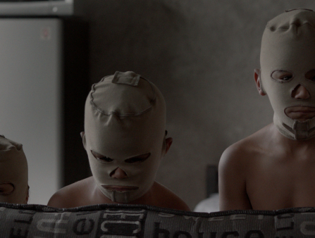 Three young shirtless boys don skin-colored masks that only reveal their eyes mouth and nose