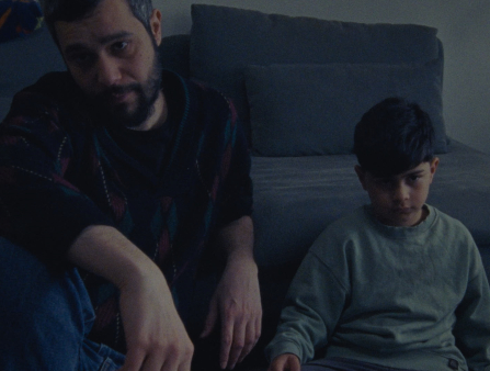 Darkly-lit shot of a middle-aged man with short grey and black hair and a beard in a sweater and jeans sitting on the floor with a young boy in a long-sleeved shirt next to him