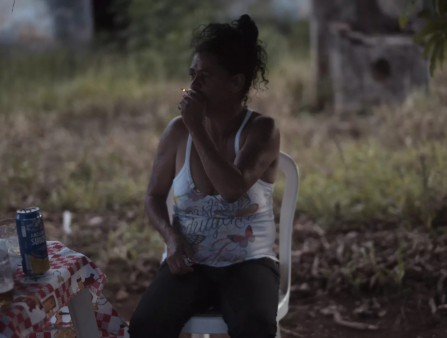 A dark skinned woman with dark curly hair in a messy bun wearing jeans and a ratty tank top sits on a plastic chair outside holds a lit cigarette up to her lips