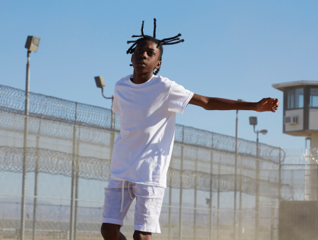 A young Black boy with dreads wearing a white tee and white shorts is seen jumping mid-air against a prison fence