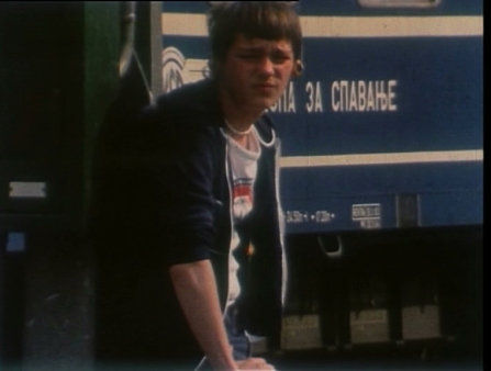 A disaffected youth in a tee and sweatshirt furrows his brow at the camera in front of a train