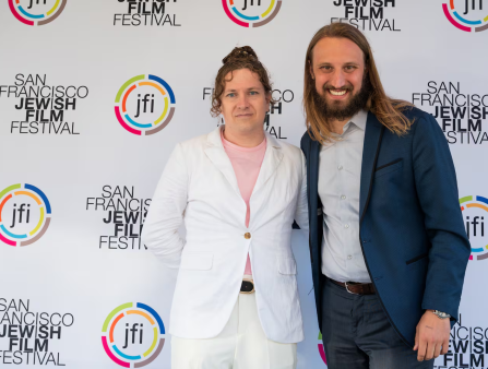 Two smiling people, one woman in a white suit and a man with long hair and a blue sportcoat, stand in front of a background which reads "jfi" and "SAN FRANCISCO JEWISH FILM FESTIVAL"