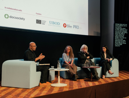 A bald man with glasses sits on an armchair. Two women and one man sit on a couch across from him. They are all on a stage with a large screen behind them