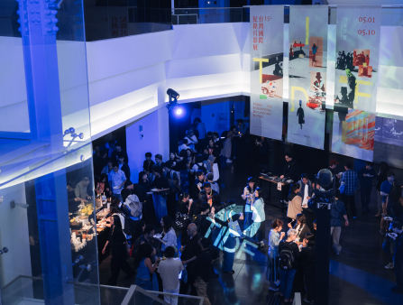 Shot from above of dozens of formally dressed people gathered in a large exhibition space. A table serves a range of food. A man uses a DJ board on a table. The room is mostly lit in indigo. Three large banners hang in the space which read "Taiwan International Documentary Festival 2026"
