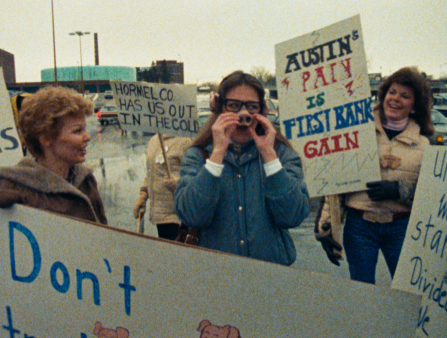 Four white women in jackets hold large signs in a parking lot. One sign reads "HORMEL CO. HAS US OUT IN THE COLD." Another reads "AUSTIN'S PAIN IS FIRST BANK GAIN." The woman in front holds a pig's snout over her nose and wears pig ears