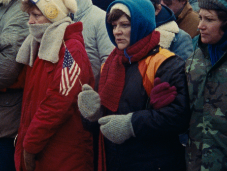 Three women in winter coats, scarves, gloves and hats lock arms in front of what appears to be a large crowd. The woman in the center holds an American flag