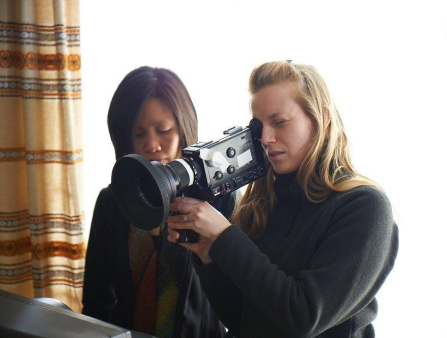 A young blond female filmmaker holds a camera against a bright window behind her; a young Asian woman with dark hair stands behind her