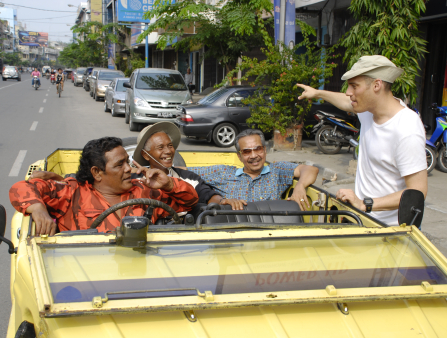 A white man in a white tee points at something behind him as three brown-skinned smiling men in a fancy yellow car interact with him out on the streets