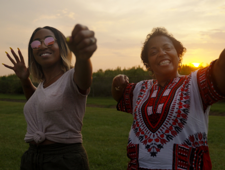 Two women appear to be drawing invisible bows and smiling in a field as the sun sets behind them. The woman on the left is younger and wears a T-shirt tied in front of her and sunglasses