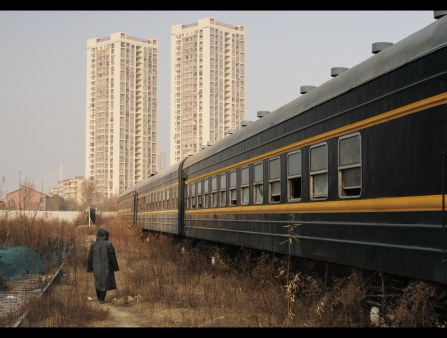 A figure in a long, hooded coat walks through overgrown vegetation alongside a parked train. In the background rise two white apartment buildings