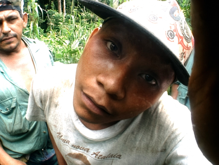 A man in a cap stares directly into the camera. The angle is distorted, as if shot with a fish eye lens. Another man in the background also looks into the camera. The two are in a densely vegetated area