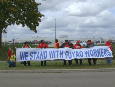 A row of 14 men and women in red stand in front of a parking lot, holding a large white banner which reads "WE STAND WITH FUYAO WORKERS" in blue text