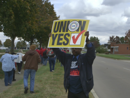 A Black man holding a yellow sign above his head, which reads "UNION YES," stands behind a crowd of marchers