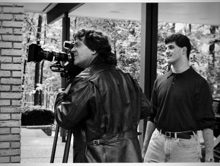On the left, a man in a black leather jacket peers into a video camera. On the right, a younger man in a tucked polo observes.  Black and white photo