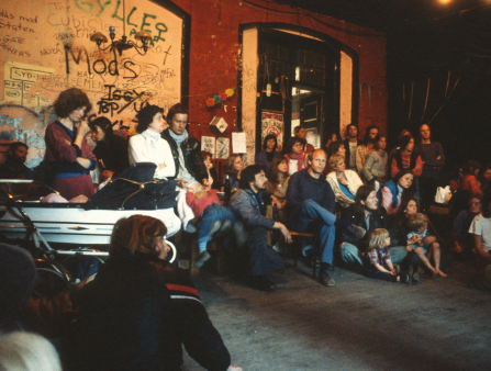 A crowd of roughly 40 people of varying ages, dressed in warm clothing, gather round in a decrepit room with graffitied walls. Most sit, some in the back stand