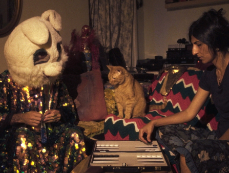 A figure in a colorful, sequined robe and bunny mask and a woman in a black shirt play backgammon. An orange cat sits between them, appearing to stare incredulously at the masked figure