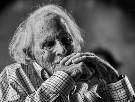 Black and white portrait of an elderly white man with mid-length white hair and a plaid collared shirt holding hands in front of face as if in contemplation