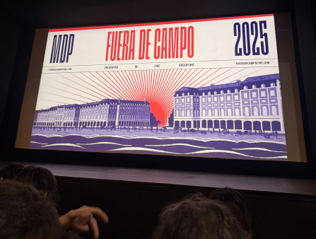 Shot of movie screen featuring the artwork for the 2025 edition of Fuera de Campo, which features a shot of government buildings outlined in blue, with bright red rays of sunshine behind them