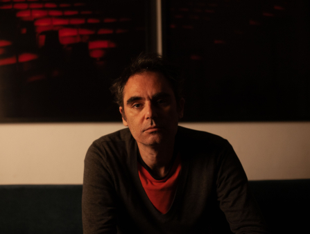Moody shot in warm light of a light-skinned man in a red tee and brown sweater looking straight at the camera