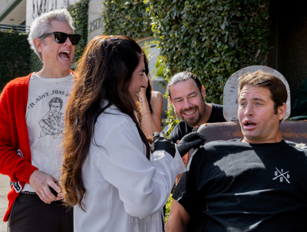 A man in a black T-shirt with comically large lips makes a pained expression. Two men and two women surround him, laughing