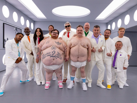 Two large men wear nothing but sneakers and diapers. One is decorated in tattoos, including a skull and crossbones and text which reads "BORN 2 DIE." Nine men and one woman stand behind the two in matching white suits. The ensemble appears in a sterile, grey room with futuristic, circular white lights