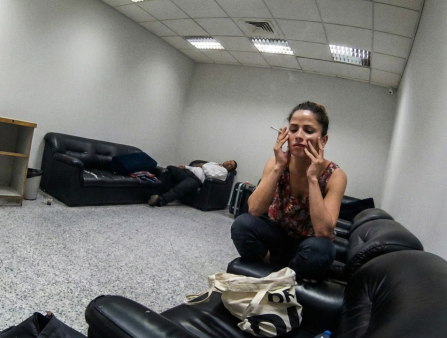 A young woman holds her head in her hands and a lit cigarette between the fingers of her right hand. She is perched on a black leather couch in a barren, grey room. More black couches appear in the background, and a man in black pants and a white shirt sleeps on one