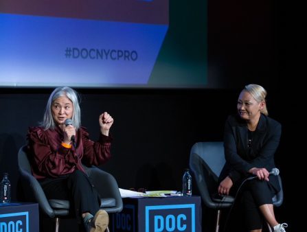 Two women sit on chairs in front of a large screen. The woman on the left holds the microphone and raises her fist. She is wearing a maroon collared shirt. Two tables on either side of her read "DOC NYC PRO"