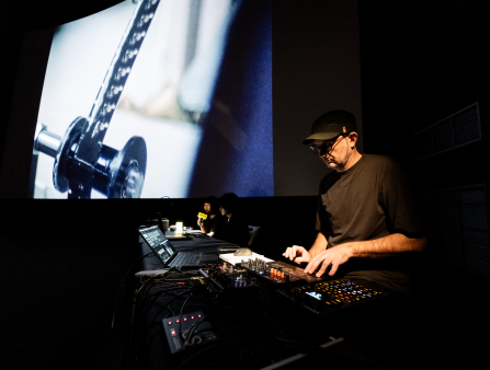 A man in a black shirt and hat, illuminated by a dim, warm light, mixes sound on a black table. Further down the table to his right sit two more figures, one holding a microphone. The table is perpendicular and to the side of a large screen, displaying a piece of metal