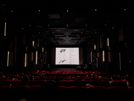 An image of a large, dark theater with red seats taken from the rear of the theater. On screen appears Chinese characters in calligraphy and a list of credits in red Chinese characters