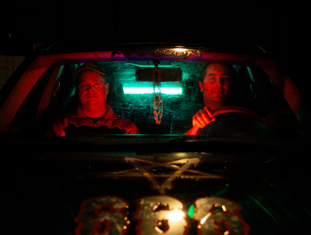 Two men sit in the cab of a truck, illuminated in red light from the vehicle's interior. The numbers 666 and an upside down pentagram appear on the hood in gold. The word "MANSON" is written in a decal at the top of the windshield