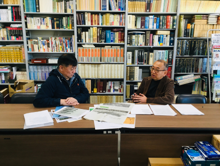 Two Asian men sit behind desk with an array of documents spread in front of them and a crowded wall of books behind them. The man on the left wears a black jacket while the man on the right wears glasses and a brown coat