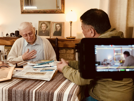 An old white man in a blue collared shirt and white cardigan places his hand on his chest while viewing a stack of books presented by an Asian man in a yellow jacket facing him from the right of the frame. The men appear on the display screen of a camera visible in the right of the frame. On the mantle behind the men are two framed portraits