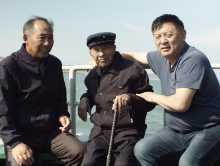 Three Asian men sit on the edge of a ship with the ocean behind them. The man on the left wears a black coat, the man in the center looks particularly aged with a hat and cane, and the man on the right wears jeans and a blue polo