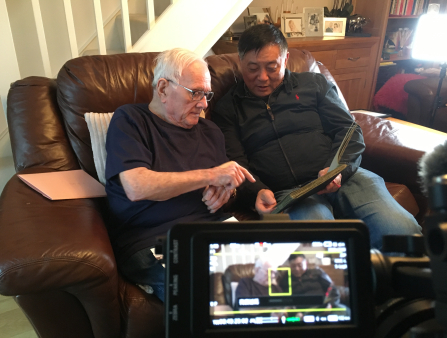And old white man and an Asian man sit on a brown leather couch looking at an album. The display screen of a camera capturing the two mean appears in the lower right of the frame