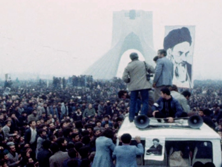 Three men with megaphones and a large poster of a man in an amameh stand atop a white van addressing a massive crowd. A large structure is visible in the far background