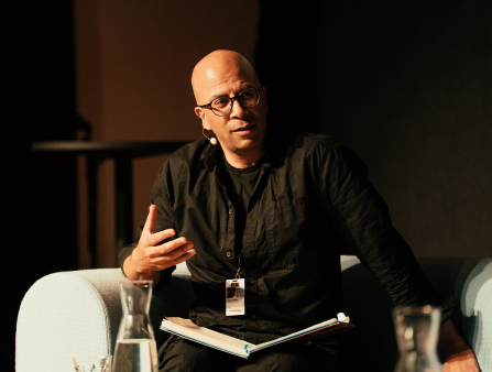 Bald man in glasses and dark brown collared shirt sits in white arm chair, gesturing with hand