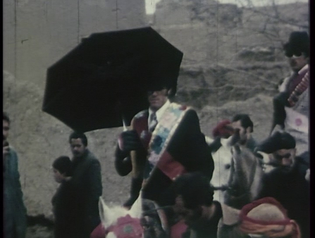 Man in suit and colored sash atop white horse holds black umbrella, another man in suit atop white horse follows him as they weave through a crowd