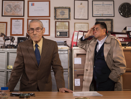 Two men appear in front of two large cabinets and a wall covered in framed documents. The man on the left is older. Staring directly into the camera, he is wearing spectacles and a brown suit with a yellow shirt and blue tie. His head is shaved. The man on the right looks up and rests his head on his hand. He wears a trenchcoat over a white button-down shirt and black cardigan.