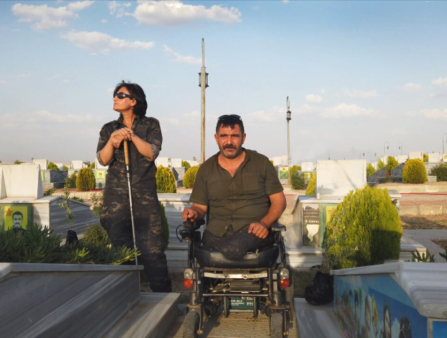 A man in a green collared shirt sits in a wheelchair. To his right (viewer's left) is a woman in sunglasses, holding a cane and looking up and off camera. The two are on a rooftop, with a blue sky and scattered cloud cover behind them
