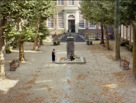 A woman sits in a sandbox in the center of a courtyard, talking to a woman standing next to her. The courtyard contains two rows of trees and leaves are scattered across the stone floor