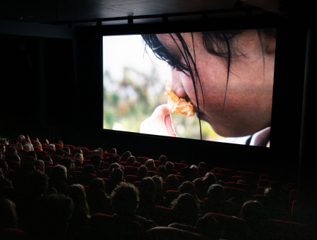 A crowd of people sit in cinema seats. The room is dark and on the screen there is an image of a young girl sniffing a flower