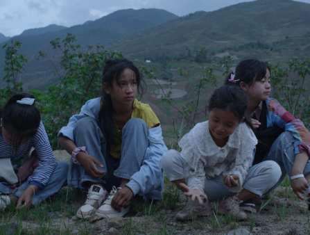 Four young Asian girls in colorful clothing sit in the grass. Rolling, grassy hills appear behind the group