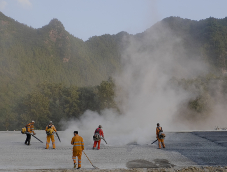 Four men in bright yellow jumpsuits and one in a bright orange jumpsuit blow a large cloud of grey dust into the air with machines they wear on their backs resembling leafblowers. One of the men in yellow holds a shovel instead. The men are in a gravel field, and large green hills rise in the background