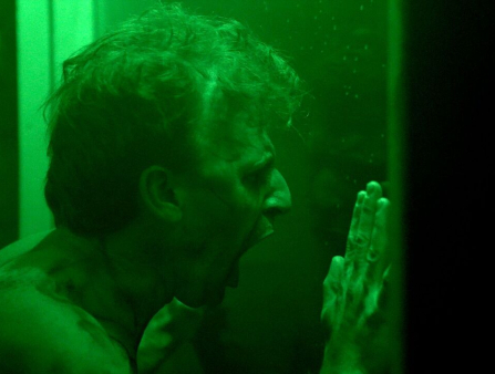 A white man presses against glass and screams, lit in bright green